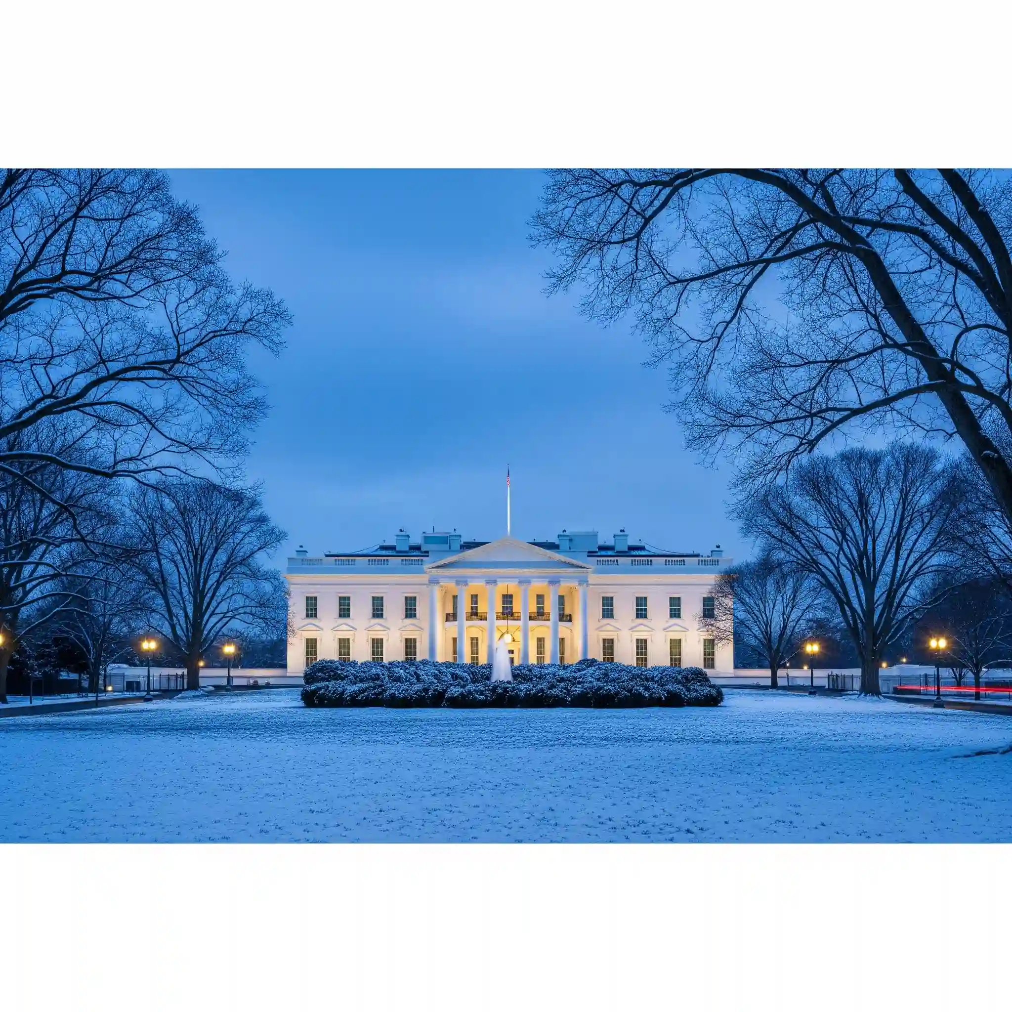 The White House at night in winter, with snow on the lawn and bare trees under a deep-blue twilight sky.